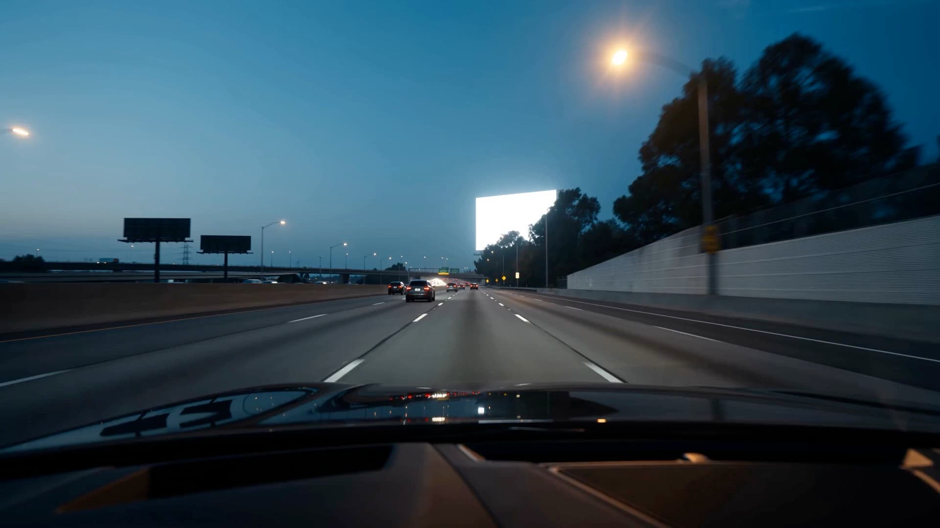 Driver's view of a freeway at dusk. One lit billboard stands on the horizon, a roadside tree cutting into its face.